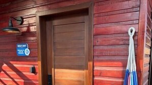 Una cabaña de madera con una puerta cerrada, un felpudo y una hamaca colgada a la derecha.