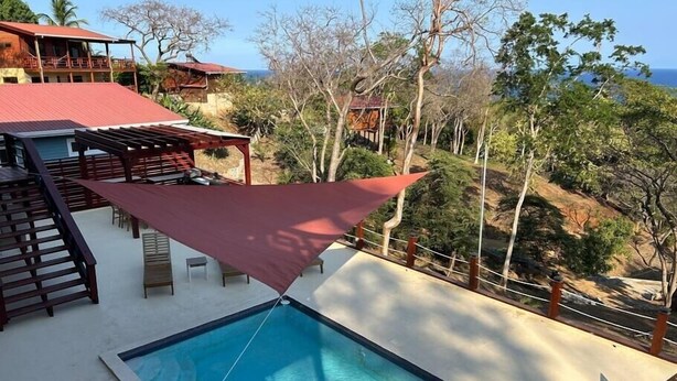 Una piscina con una vela parasol roja, sillas de madera en la terraza y vistas al océano y las colinas.