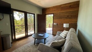 Una sala de estar moderna con un sofá, una pequeña mesa redonda y un televisor. Amplios ventanales corredizos dan a una terraza exterior.