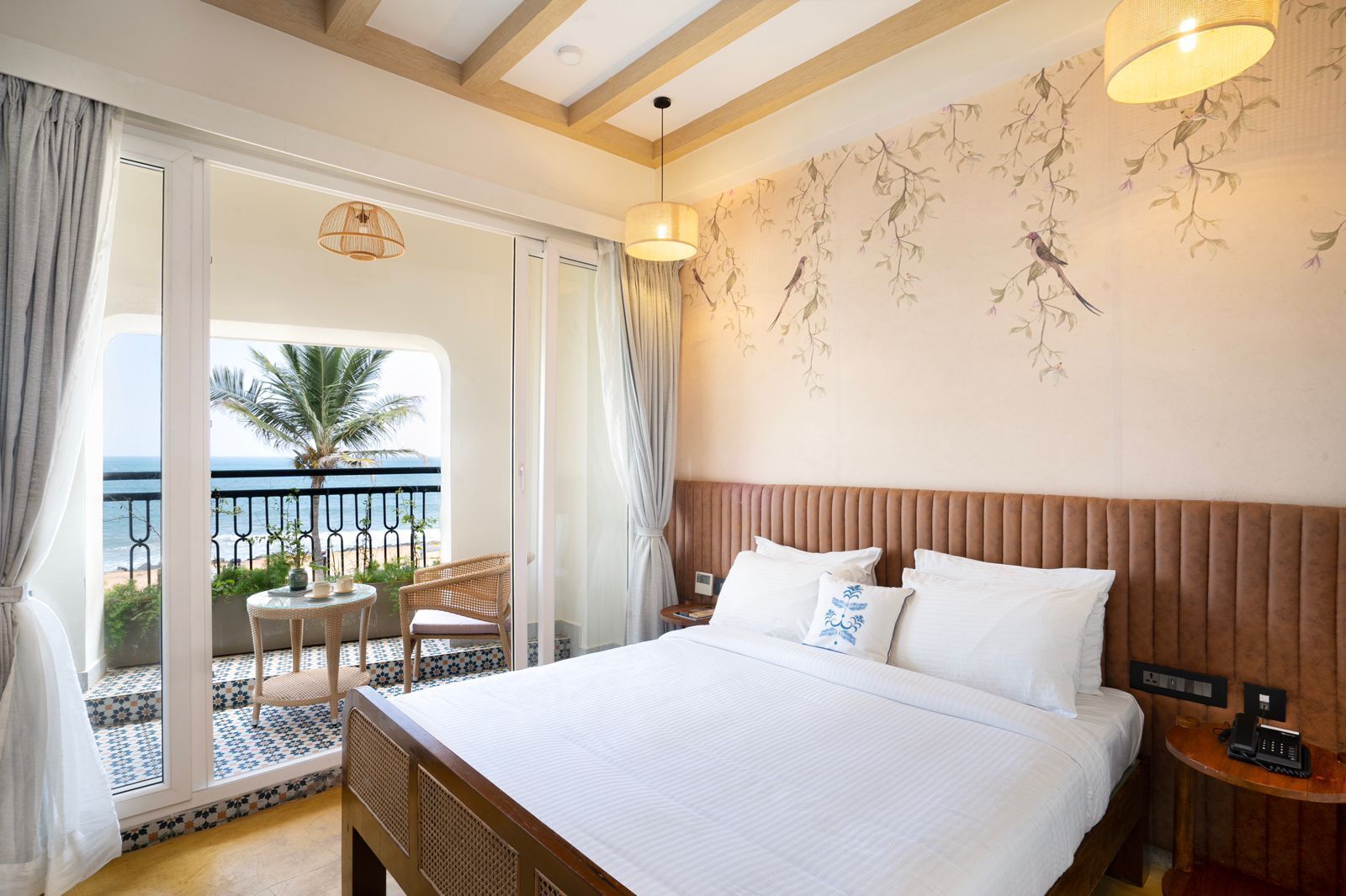 A hotel room with a large bed, a balcony with a view of the sea, and a small table with chairs.