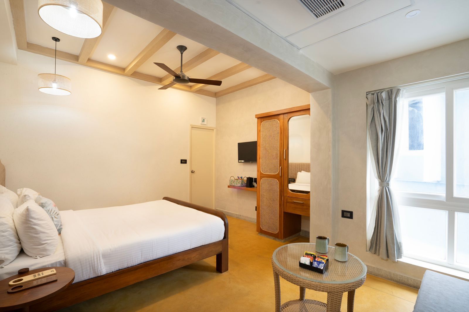 Une chambre d’hôtel comprenant un lit, un ventilateur de plafond, une télévision, une armoire, une petite table avec quelques objets, et une fenêtre avec des rideaux.