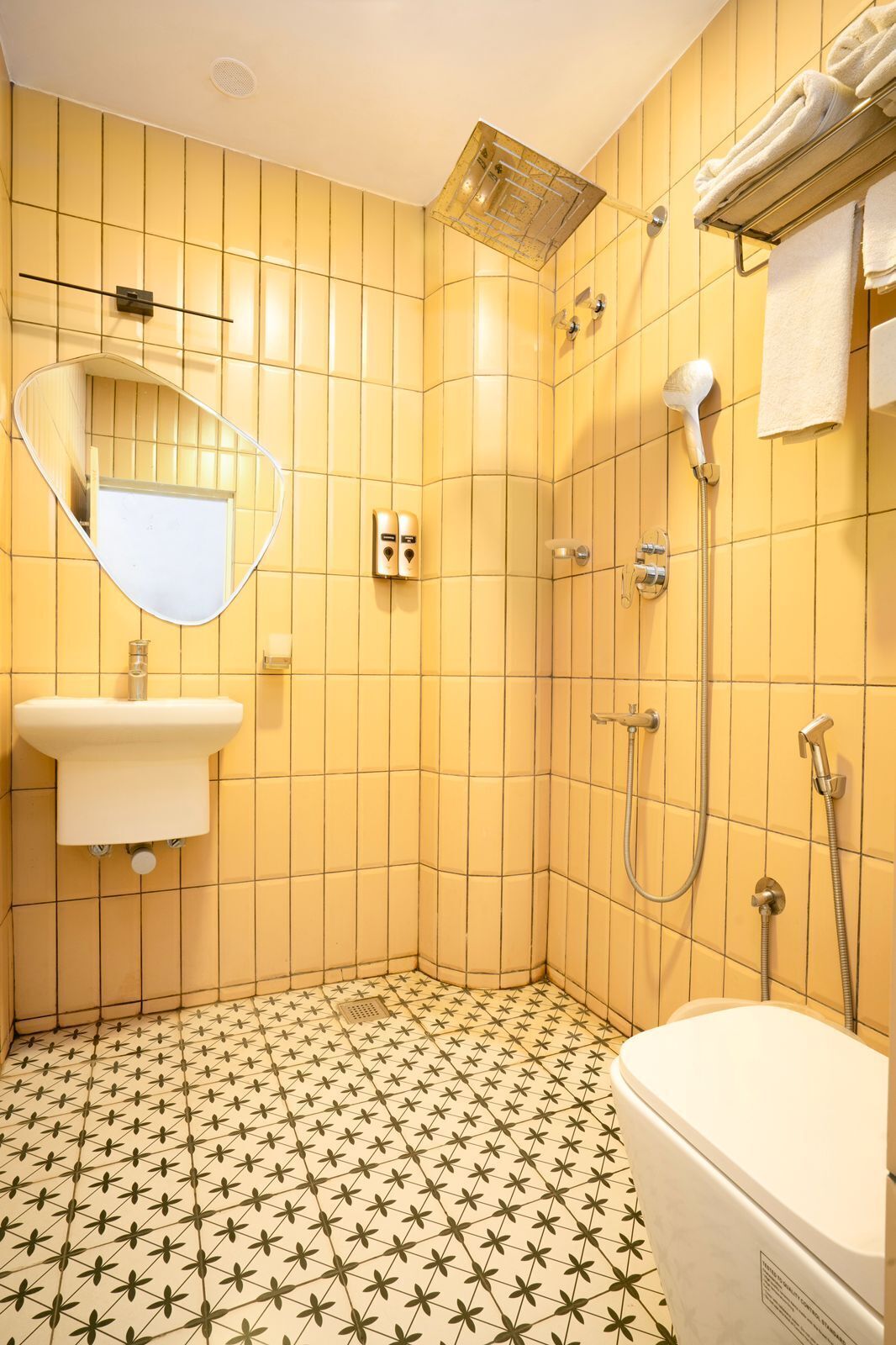 Une salle de bain avec des murs recouverts de carreaux jaunes, un lavabo blanc, un miroir, une douche avec un pommeau de douche amovible et des toilettes.