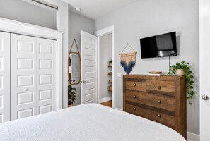 A bedroom with a bed, a wooden dresser, a TV on the wall, and a door with a mirror.