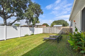 A backyard with a white fence, a tree, and a patio area with two chairs.