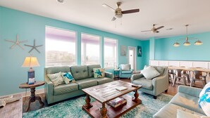A living room with teal walls, a wooden coffee table, a grey sofa, and a dining area with a table and chairs.