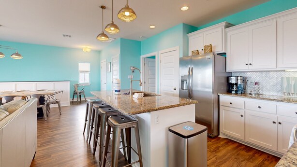 A modern kitchen with white cabinets, a granite countertop, and stainless steel appliances. There are bar stools and a dining area with a table and chairs.
