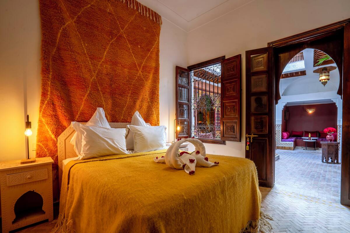 A cozy bedroom with a bed, a nightstand, a wall tapestry, and a view through an open door to a balcony.