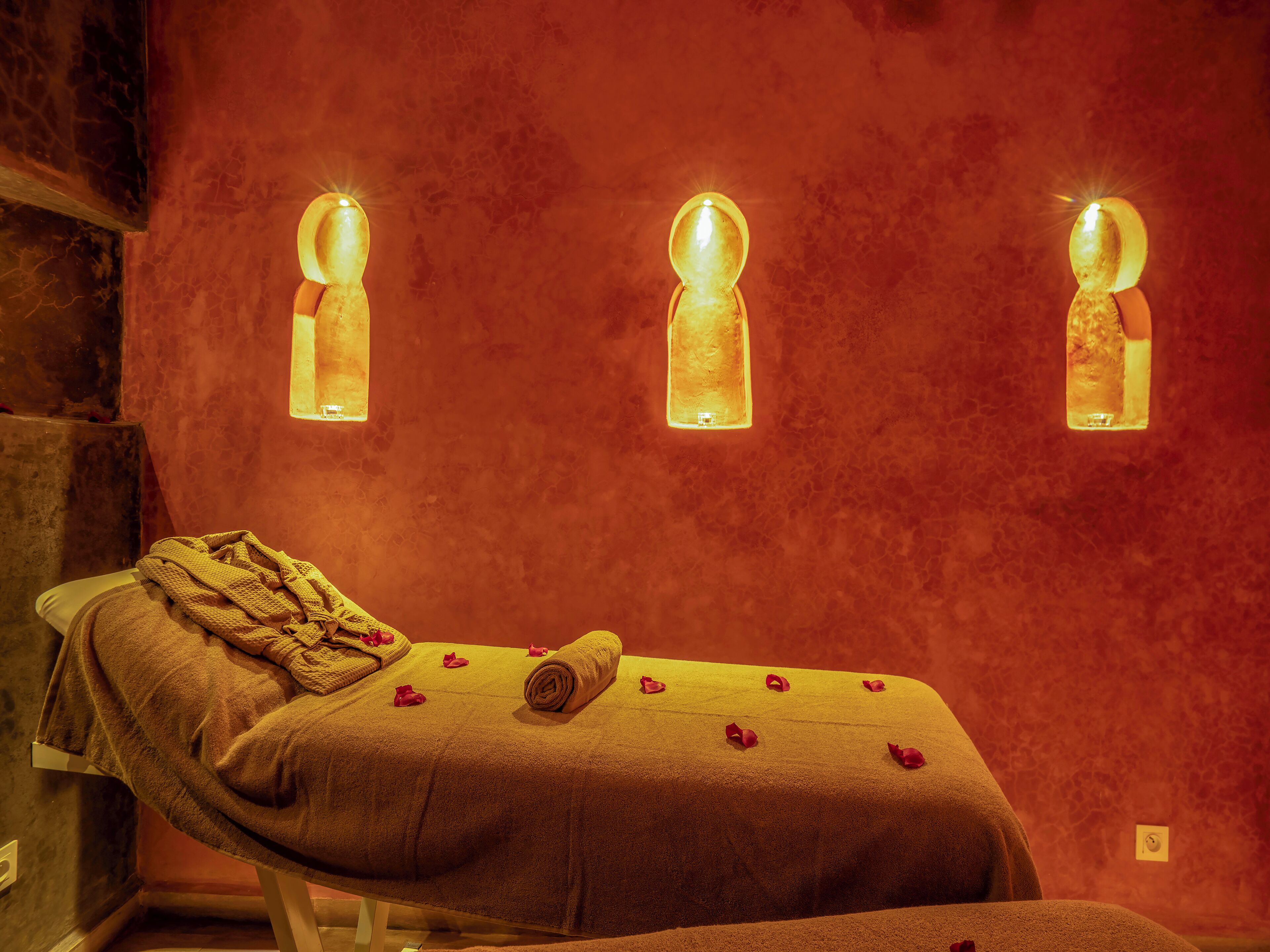 Une table de massage, agrémentée d’une serviette et de pétales de rose, est placée devant un mur rouge à texture, sur lequel sont fixées trois lampes.