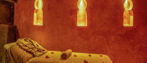 Une table de massage, agrémentée d’une serviette et de pétales de rose, est placée devant un mur rouge à texture, sur lequel sont fixées trois lampes.