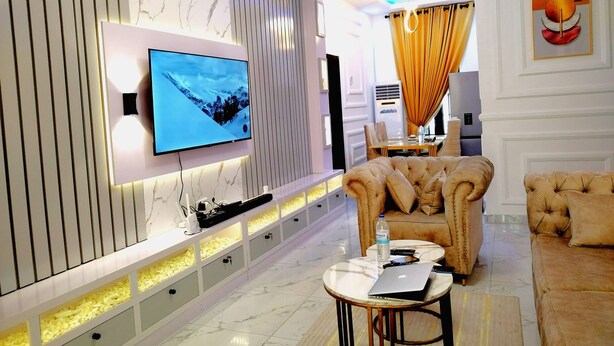 A modern living room with a flat-screen TV mounted on a wall, a plush sofa, a coffee table, and a dining area in the background. - Kings' Place Lekki - 2Bedrooms (Lekki)