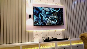 A modern living room with a flat-screen TV mounted on a marble wall, a white entertainment unit below, and a small aquarium. - Kings' Place Lekki - 2Bedrooms (Lekki)
