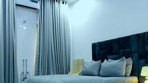 A modern bedroom with a bed, curtains, a bedside table with a lamp, and a wall-mounted air conditioning unit. - Kings' Place Lekki - 2Bedrooms (Lekki)