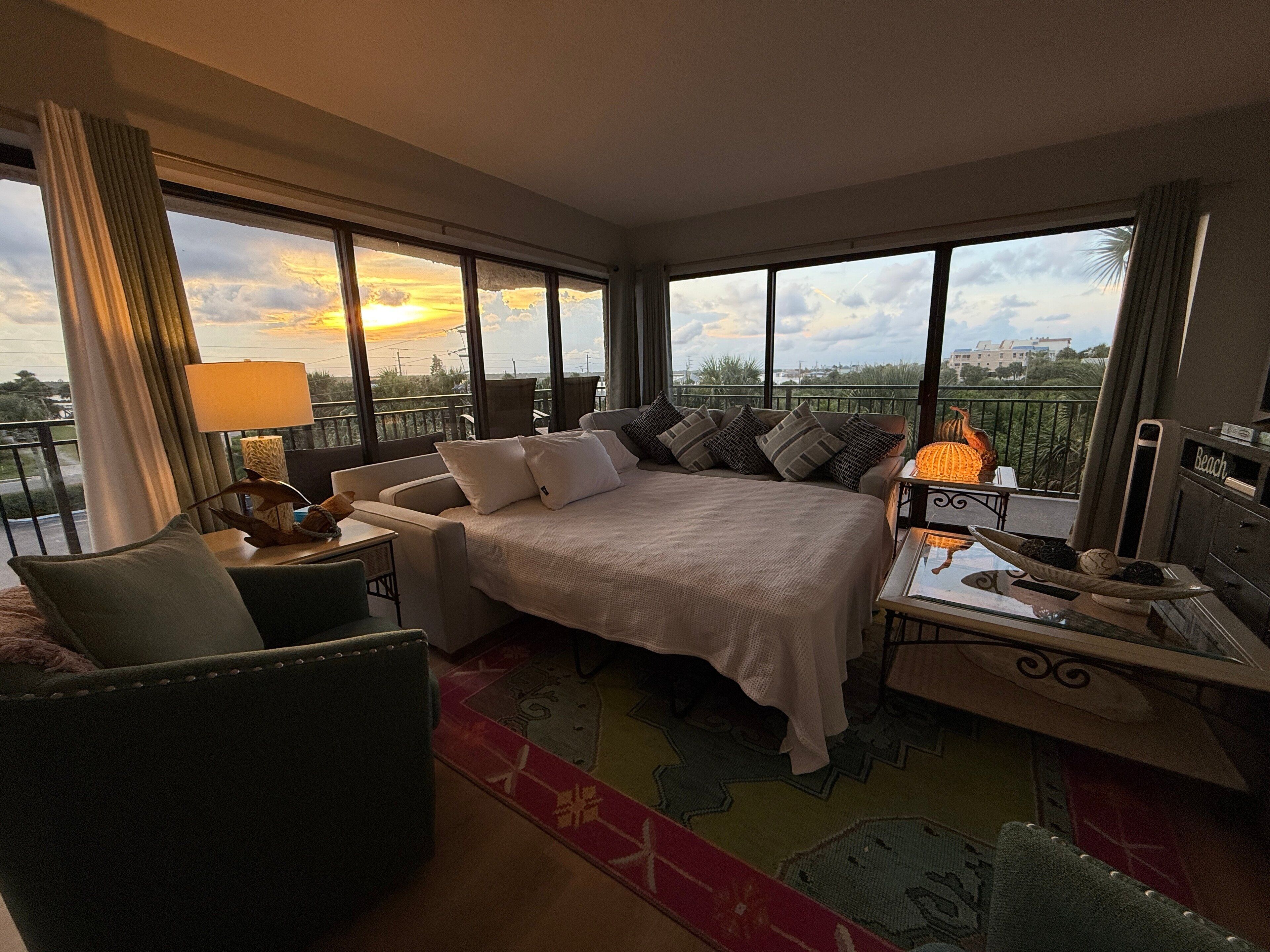 A hotel room with a bed, a desk, a chair, and a balcony with a view of the sunset.