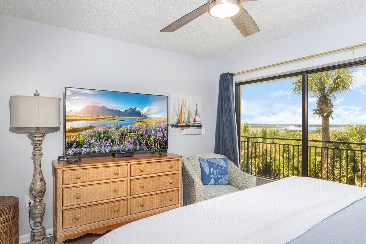 A bedroom with a bed, a dresser, a television, a lamp, and a balcony with a view of palm trees and a clear sky.
