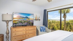 Et soveværelse med seng, kommode, fjernsyn, lampe og en balkon med udsigt til palmer og en klar himmel.