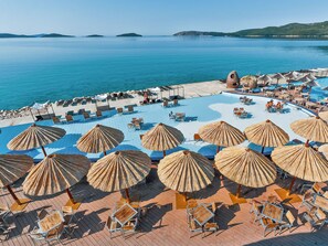 A resort pool area with sun loungers and thatched umbrellas, surrounded by a scenic view of the sea and distant islands.