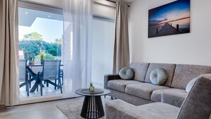 A modern living room with a grey sofa, a small round table, and a glass door leading to an outdoor seating area with a table and chairs.