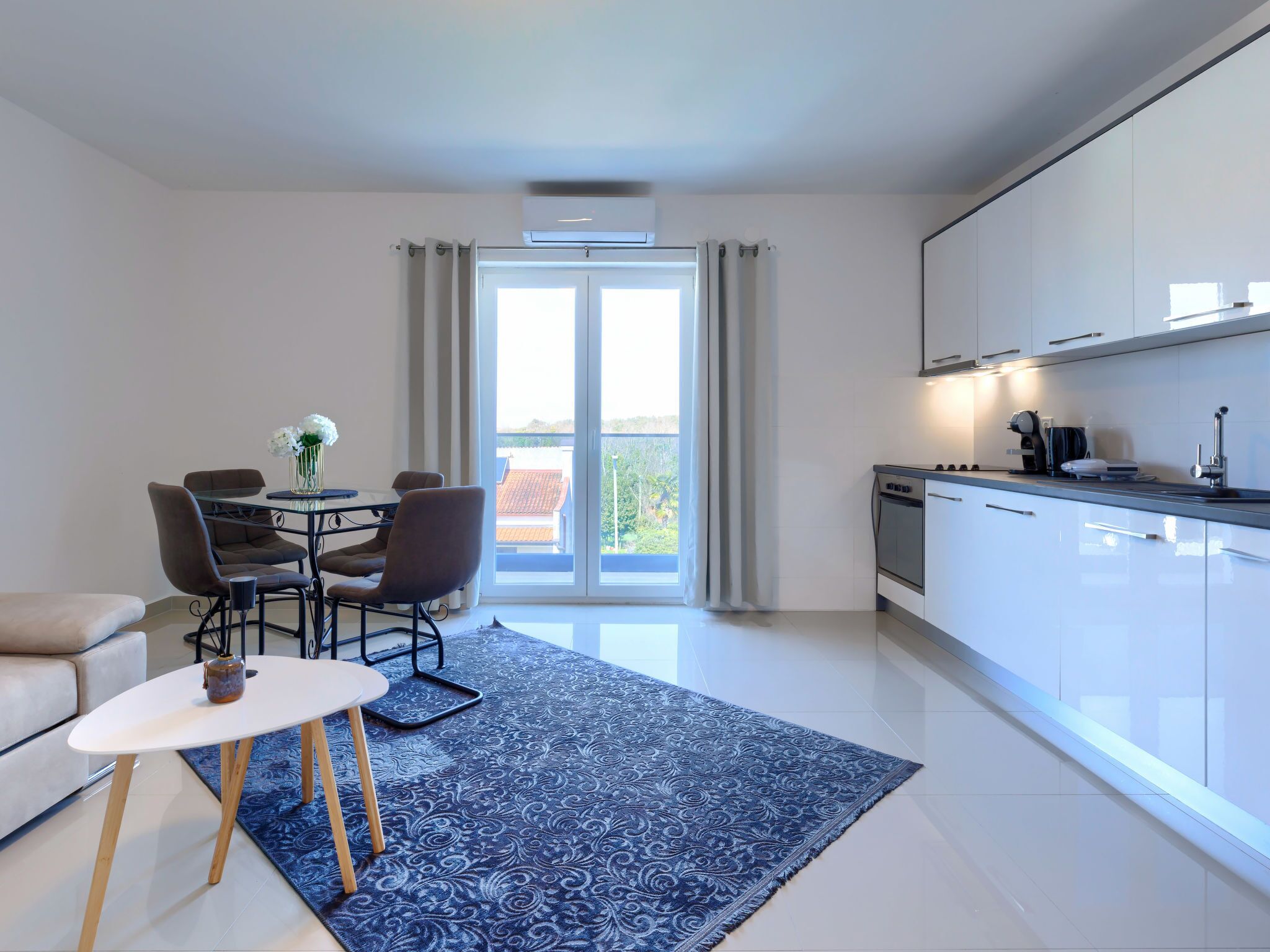 A modern living room with a dining area, a kitchen, and a large sliding glass door leading to a balcony.