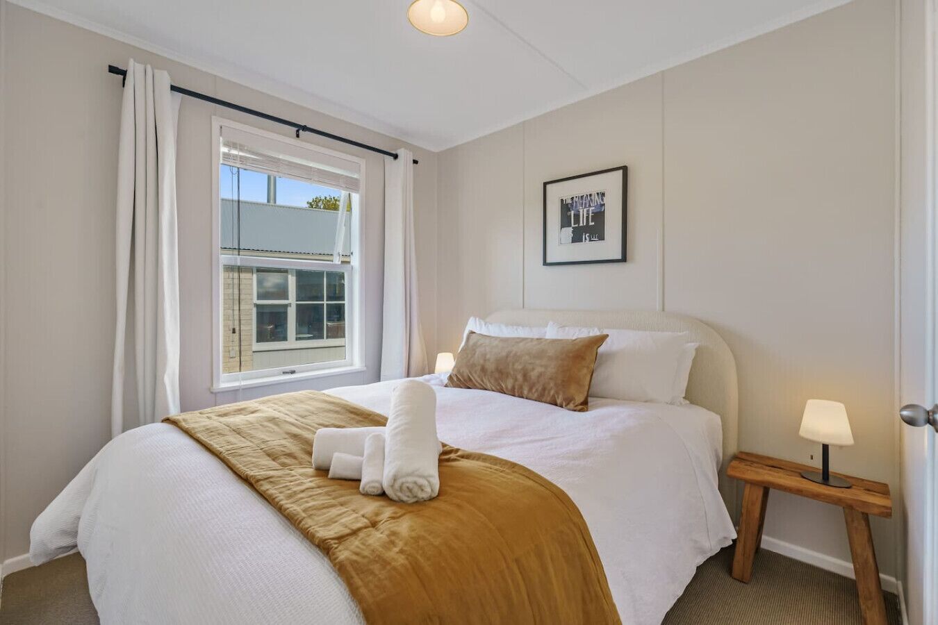 Una cama bien hecha con una manta marrón y almohadas blancas, una lámpara de noche y una ventana con cortinas.