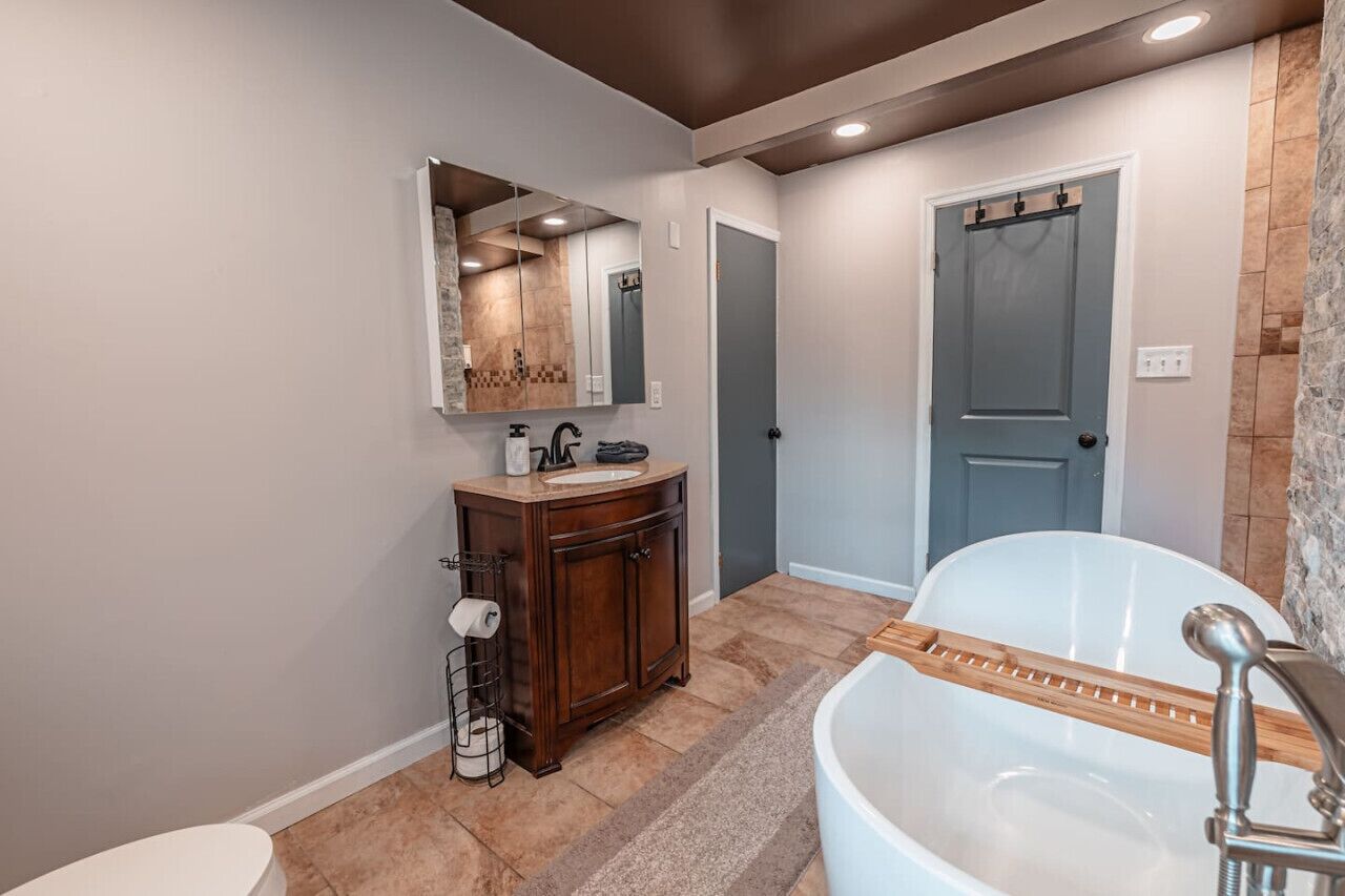 A bathroom with a freestanding bathtub, a wooden vanity, a large mirror, and a door leading to another room.