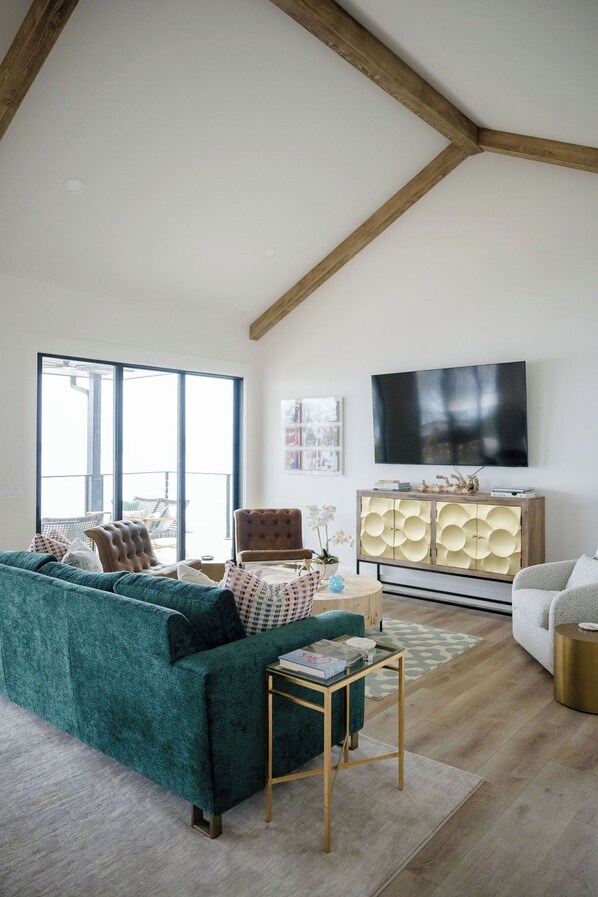 A spacious living room with a high ceiling, a large window, a green sofa, a wooden cabinet, and a flat-screen TV. - Playin Possum! NEW, Modern 5 Bed 4.5 Bath Retreat with Dock, 2 Balconies, HotTub (Graford)
