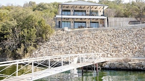 Un edificio moderno con techo plano y ventanas amplias, ubicado cerca de una masa de agua con un puente blanco que lo atraviesa.