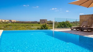 A clear blue pool with lounge chairs and an umbrella, surrounded by a view of buildings and greenery. - Kenn Gozitan villa and pool, L-Gharb Gozo, Malta (L-Gharb Gozo)