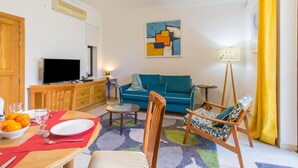 Una sala de estar con un sofá, un televisor, una mesa de comedor con un mantel individual rojo y una alfombra colorida.