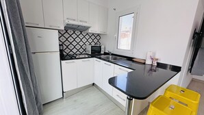 Et moderne køkken med hvide skabe, en sort bordplade og en mikroovn. Der er et vindue med udsigt til bygninger og en gul skammel.