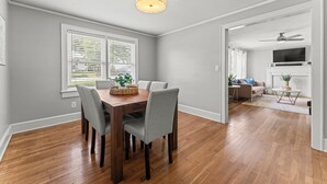 A dining area with a wooden table and chairs, a ceiling light, and a view to a living room with a sofa and TV.
