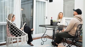 A cozy outdoor seating area with a macrame chair, a table with a plant and a bottle, and a glass of wine.