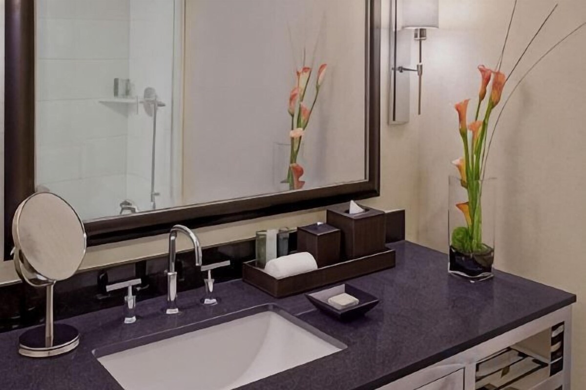 A bathroom with a large mirror, a sink, a vanity with a mirror, and a vase with flowers.