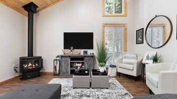 A cozy living room with a fireplace, a TV, comfortable seating, and a large window with blinds.