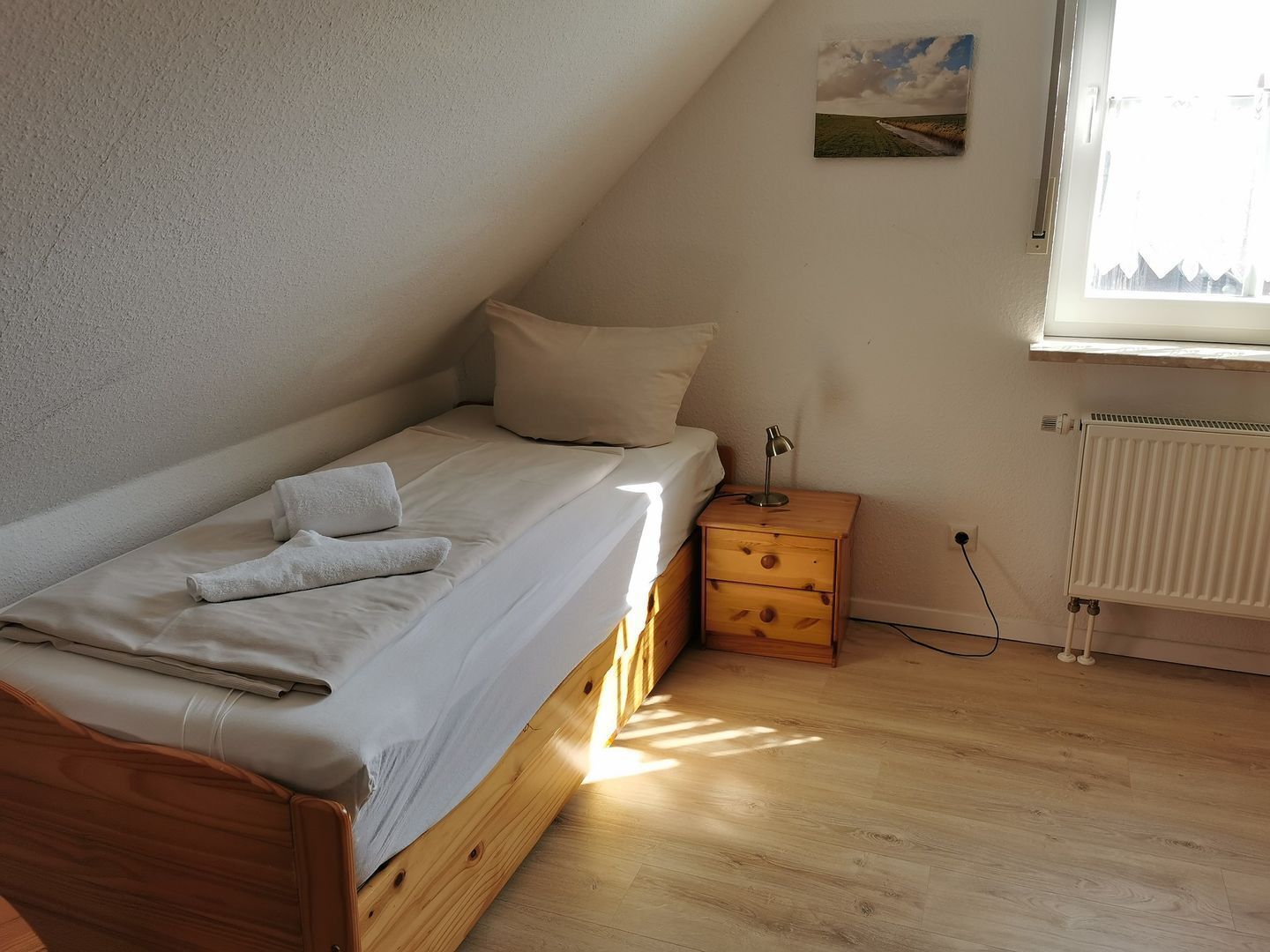 Une petite chambre avec un lit simple, un plafond en pente, une table de chevet avec une lampe, un radiateur et une fenêtre avec des rideaux.