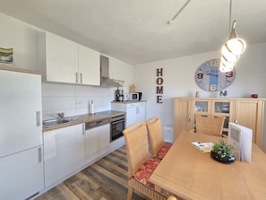 A compact kitchen with white cabinets, a wooden countertop, and a dining area with wicker chairs and a wooden table. - Ferienwohnung Hus Achtern Diek - Herzlich Willkommen in der Ferienwohnung Hus Achtern Diek! (Neßmersiel)