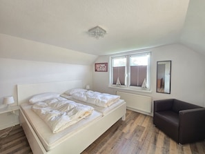 A bedroom with two beds, a window with blinds, a radiator, a chair, and a ceiling light fixture. - Ferienwohnung Hus Achtern Diek - Herzlich Willkommen in der Ferienwohnung Hus Achtern Diek! (Neßmersiel)