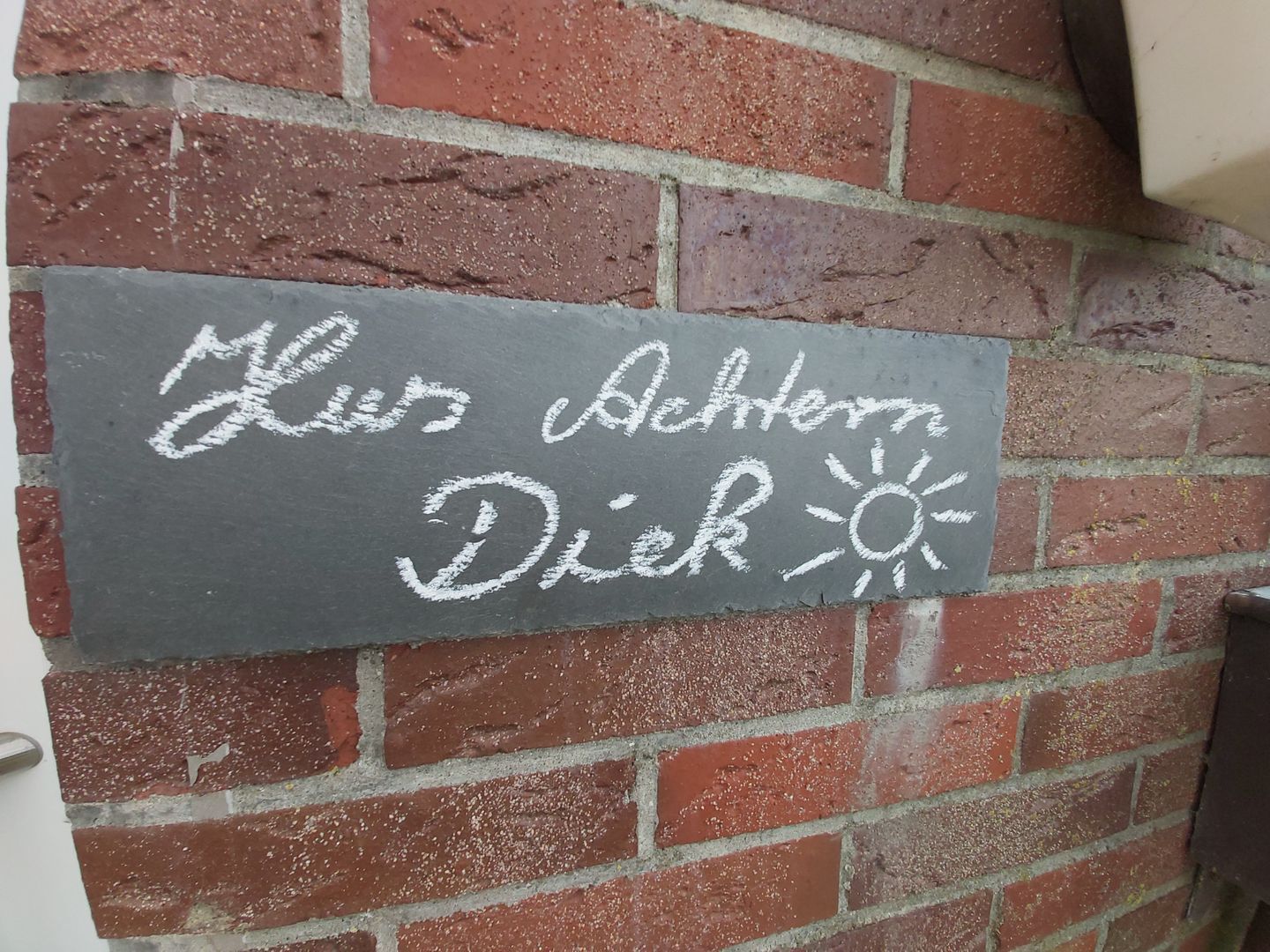 A chalkboard sign on a brick wall with the text "Mies Achtern Diek" and a sun symbol.