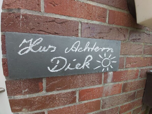 Un cartel de pizarra en una pared de ladrillo con el texto 'Mies Achtern Diek' y un símbolo de sol.