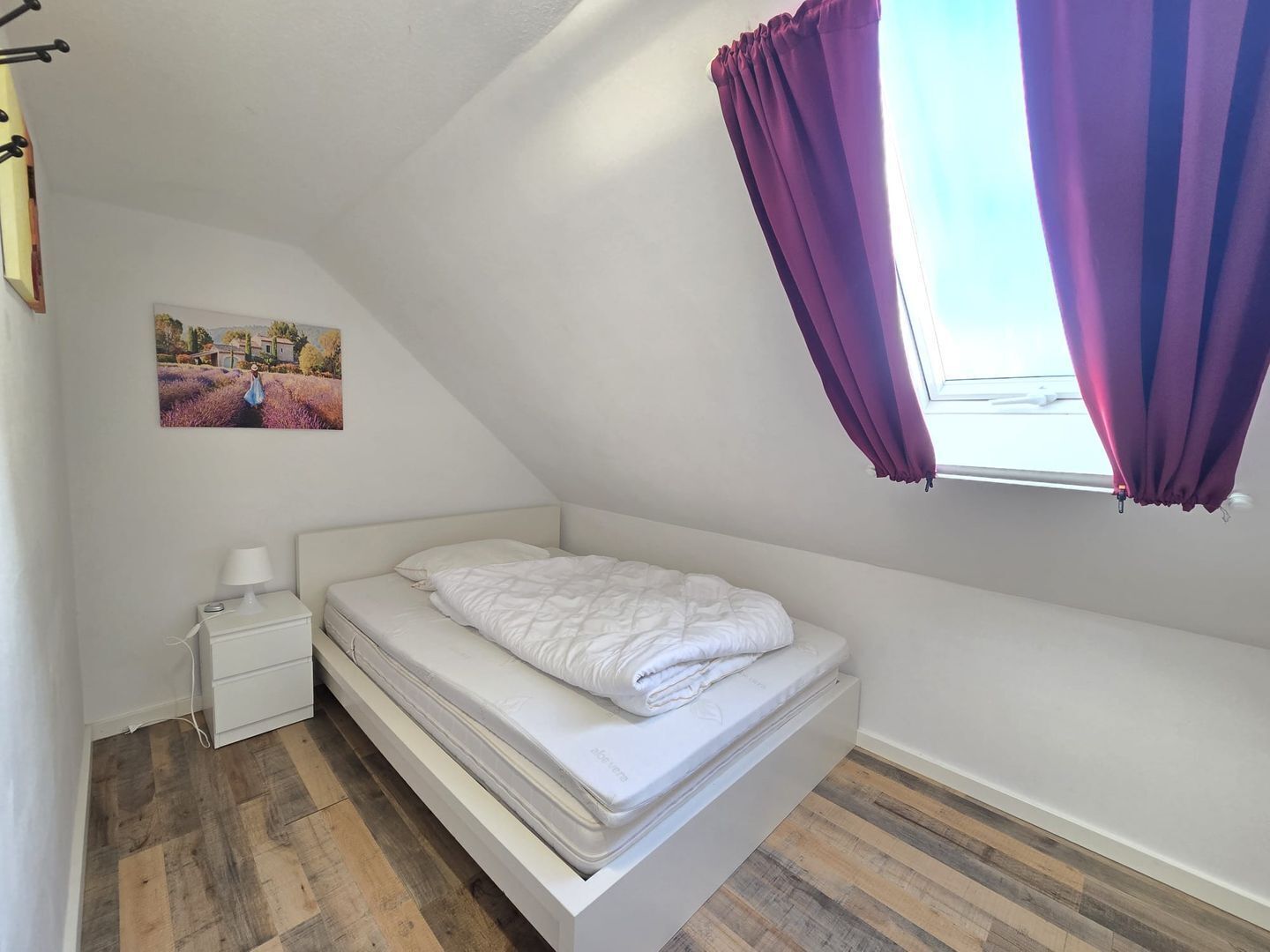Un dormitorio estilo loft pequeño y blanco con una cama individual, una mesita de noche, una lámpara, un cuadro en la pared y cortinas moradas en una ventana.