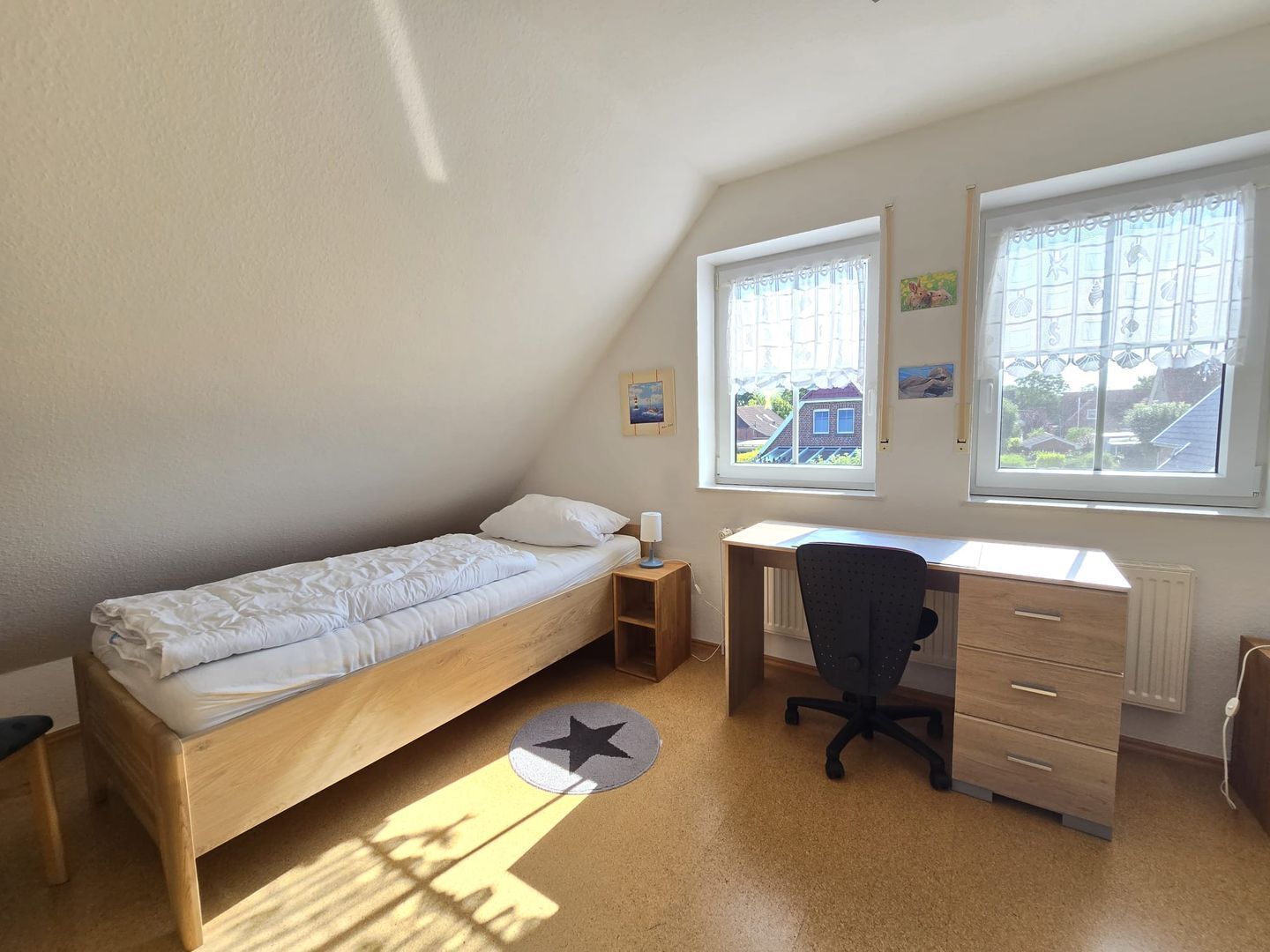 Una habitación pequeña y bien iluminada con una cama individual, un escritorio y dos ventanas.