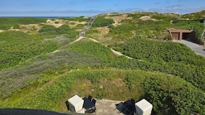 Un paisaje costero con una escalera de madera que sube, una pequeña construcción de ladrillo y una mesa con sillas al frente.