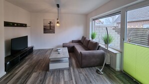 Ein modernes Wohnzimmer mit Flachbildfernseher, grauem Sofa, Couchtisch und großem Fenster mit Außenblick.