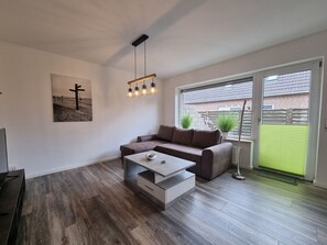Een moderne woonkamer met een hoekbank, een salontafel en een groot raam met uitzicht op een stenen huis en groen.