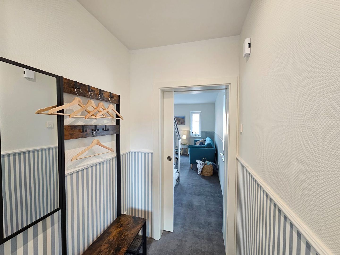 A hallway with a coat rack, a bench, and a door leading to a room with a blue sofa and a window.