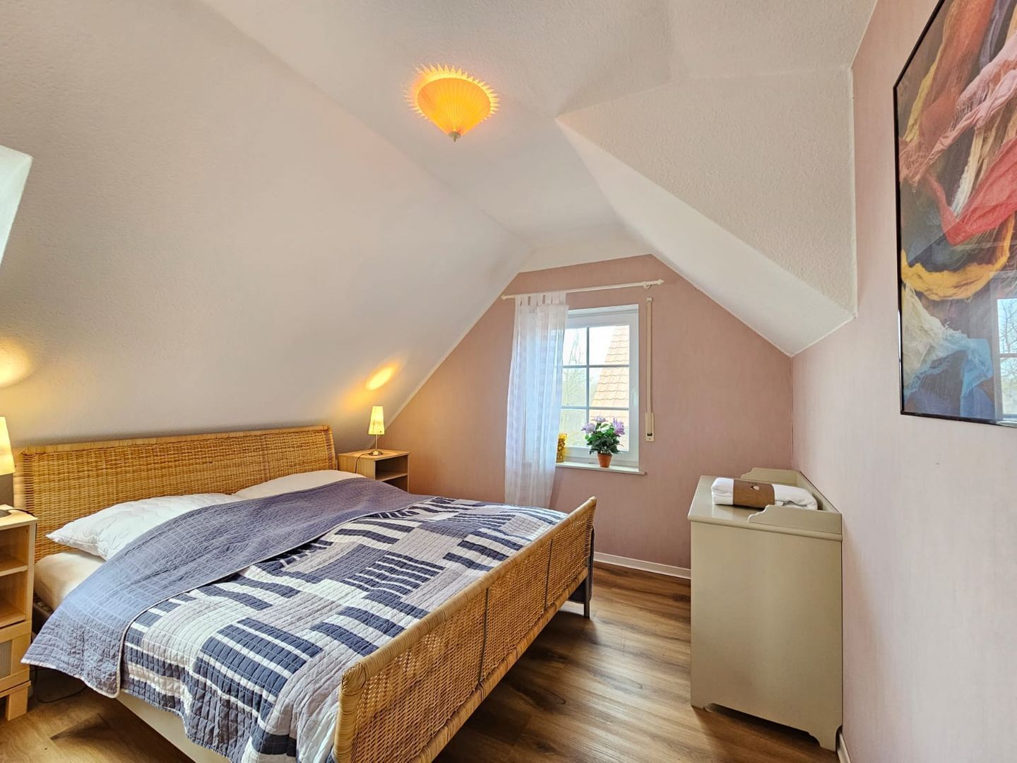 Un dormitorio acogedor con techo inclinado, cama con manta a rayas, lámpara de noche, un mueble pequeño y una ventana con cortina.