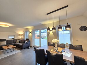A modern living room with a sofa, coffee table, and dining table with chairs. There are pendant lights above the table and a clock on the wall. - Ferienhaus Urlaubsglück - Herzlich Willkommen im "ferienhaus Urlaubsglück "! (Dornum)
