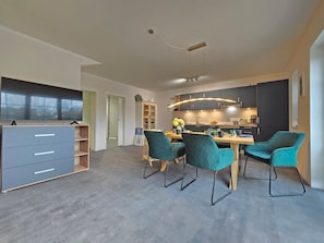 Una cocina moderna con armarios oscuros, una zona de comedor con sillas color turquesa y un televisor de pantalla plana sobre un mueble gris.
