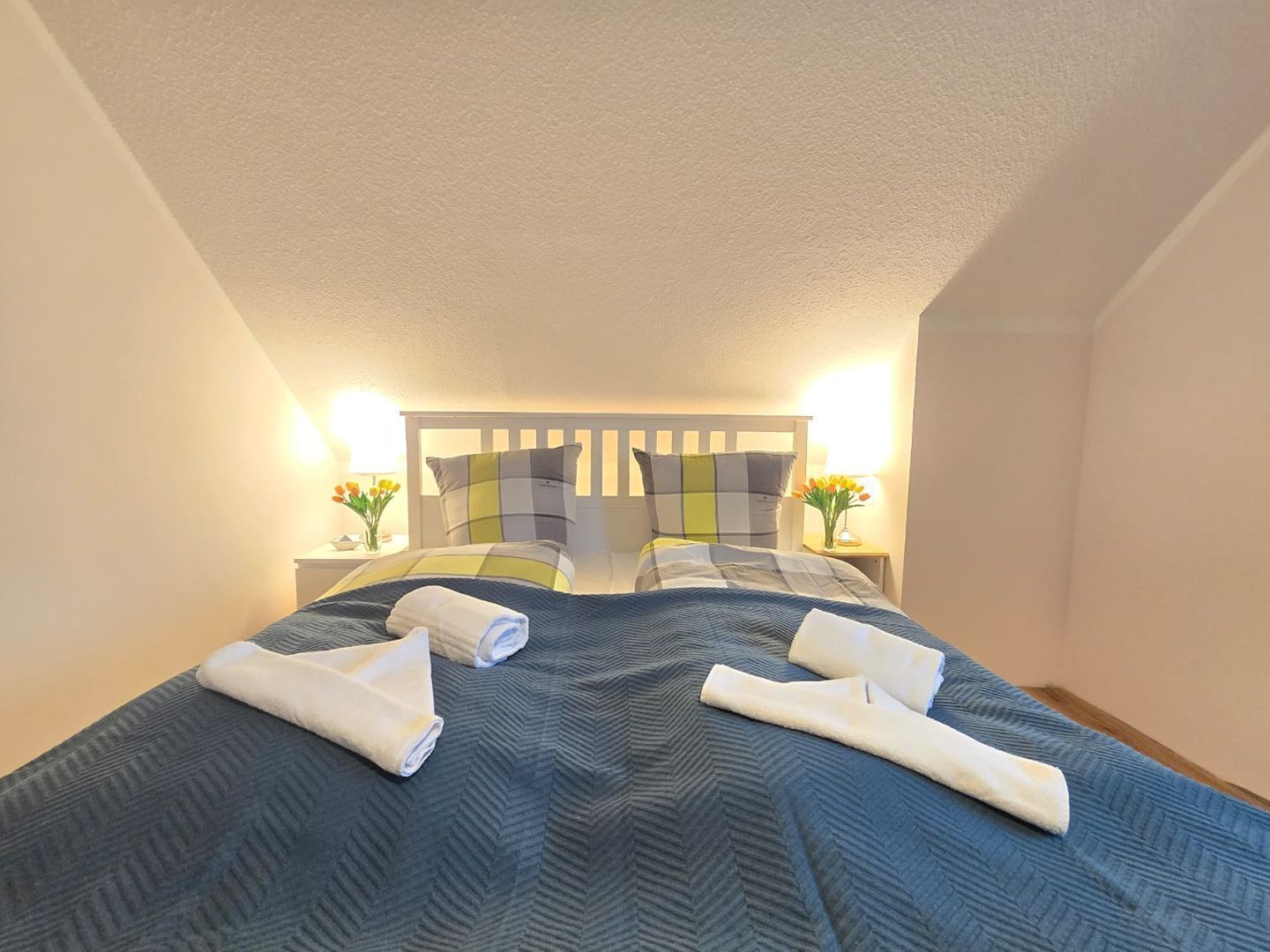 A double bed with a blue quilt, two pillows, and two rolled towels.