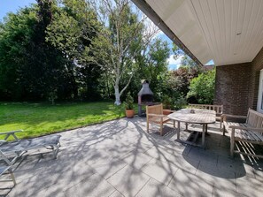 A patio area with a brick wall, a wooden table, and chairs. There is a grill and a tree in the background.
