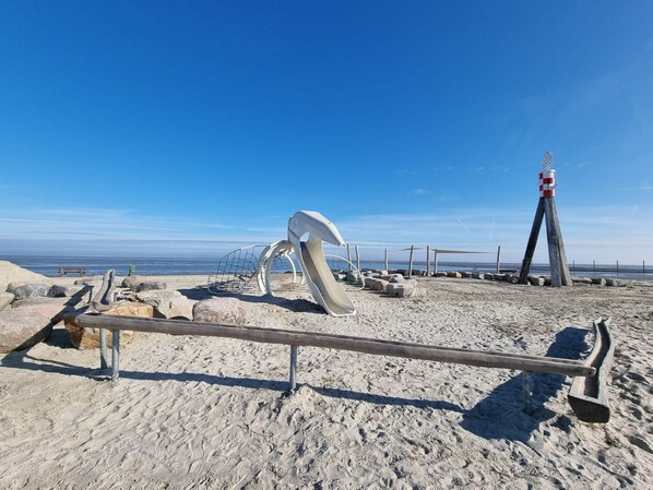 Una playa con una escultura de metal, un banco de madera y un faro al fondo.